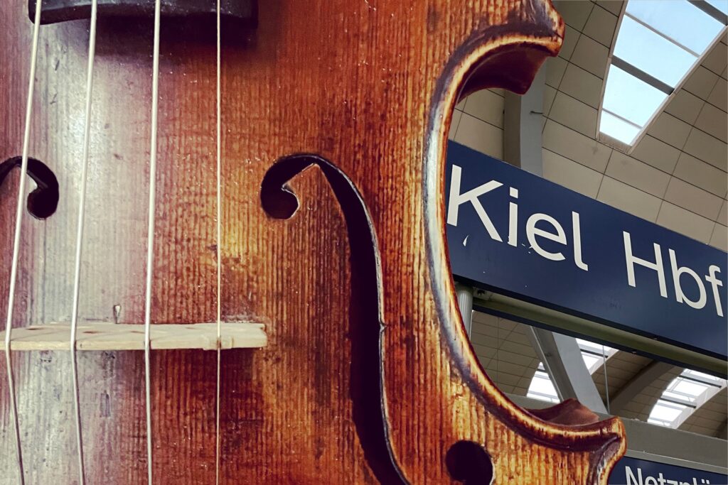 Symbolbild: Geige im Vordergrund, Bild vom Schild vom Kieler Hauptbahnhof im Hintergrund